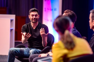 Ali Niknam sits on stage on a chair and talks to the other speakers.